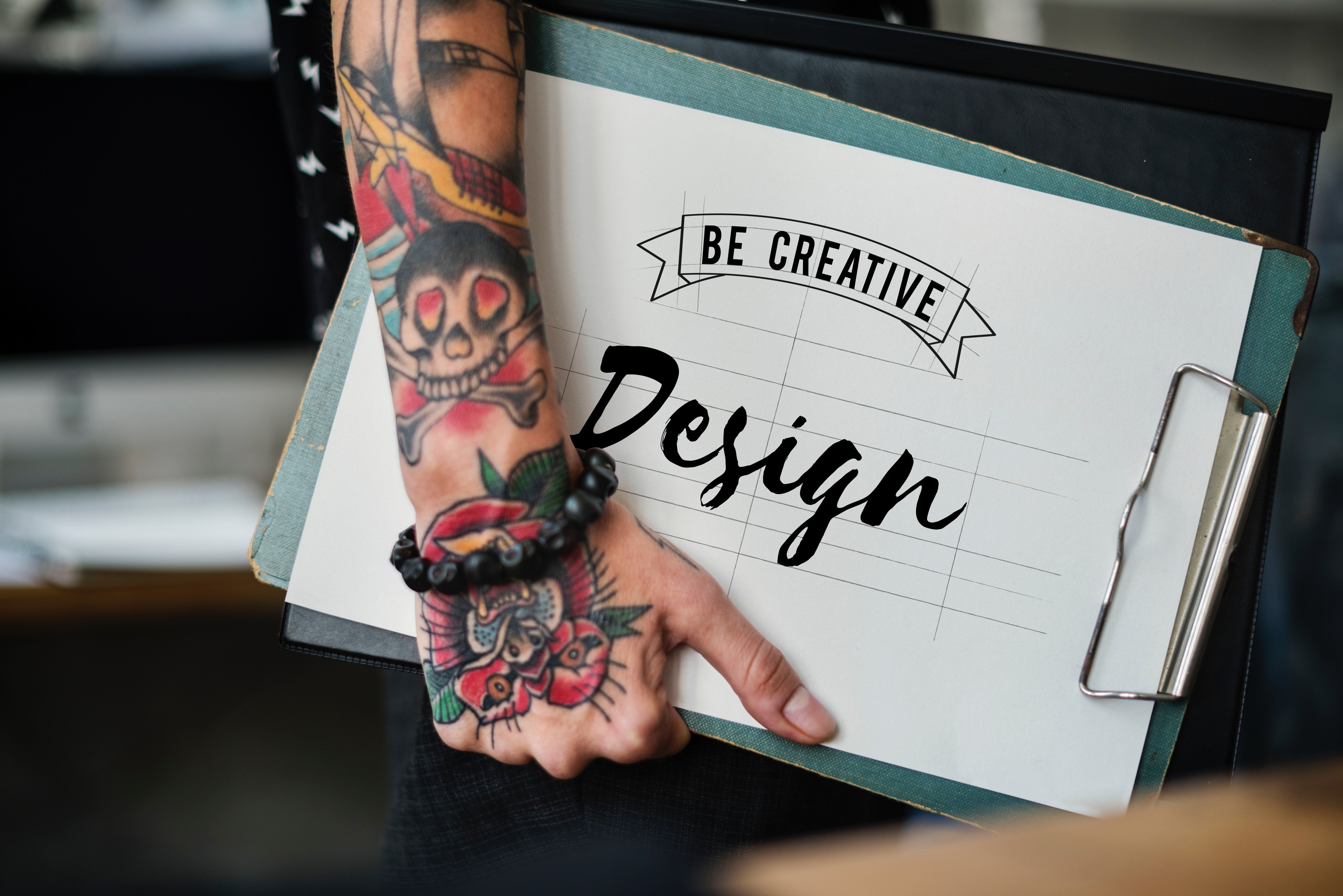 Tattooed arm holding a clip board that says Be Creative Design - New Wave Marketing Calgary